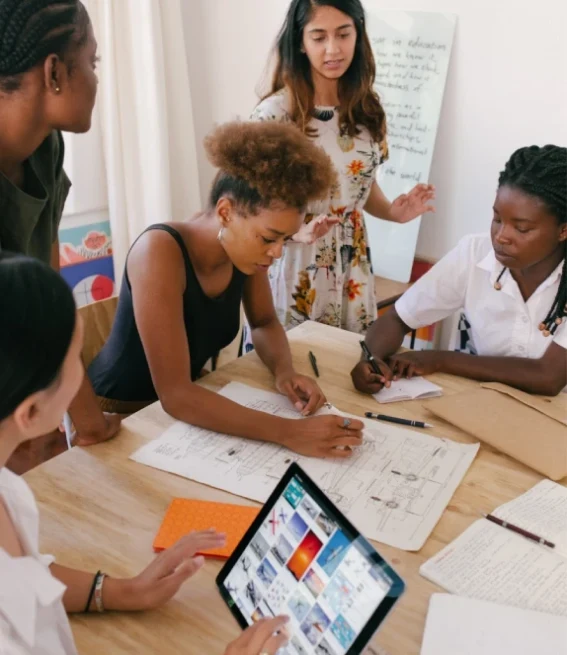 A group of five women collaborating around a table with papers, notebooks, and a tablet.