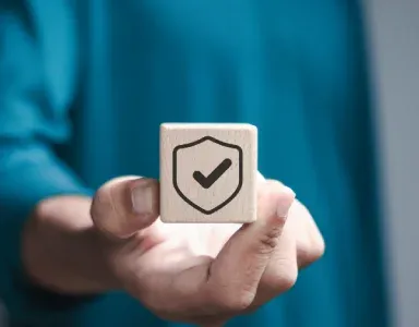 A hand holding a wooden block with a shield and checkmark, symbolizing trust.