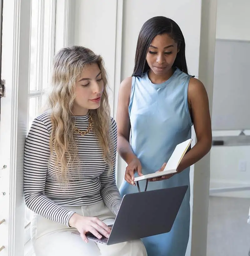 Two women working together, one with wavy blonde hair using a laptop, the other with dark hair holding a notebook.