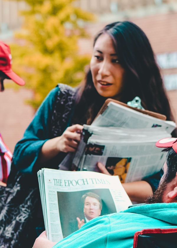 Woman holding newspapers and a clipboard, talking to a person reading "The Epoch Times" newspaper.