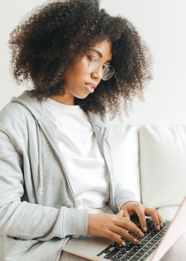 Woman with curly hair and glasses using a laptop, wearing a gray hoodie and white shirt.