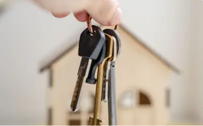 A hand holding keys with a small wooden house in the background.