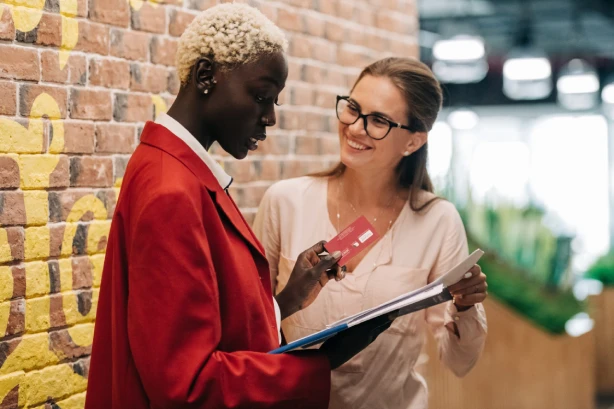 Two women talking, one holding a credit card and a notebook. One wears a red blazer, the other glasses and a light blouse.