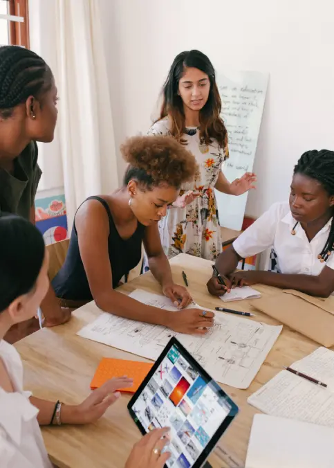 A group of five women collaborating around a table with papers, notebooks, and a tablet.