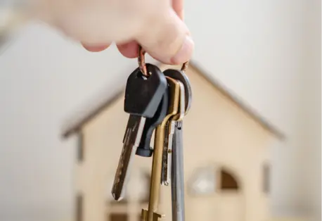 A hand holding keys with a small wooden house in the background.