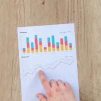 A hand pointing at a piece of paper with colorful bar and line graphs, placed on a wooden surface.