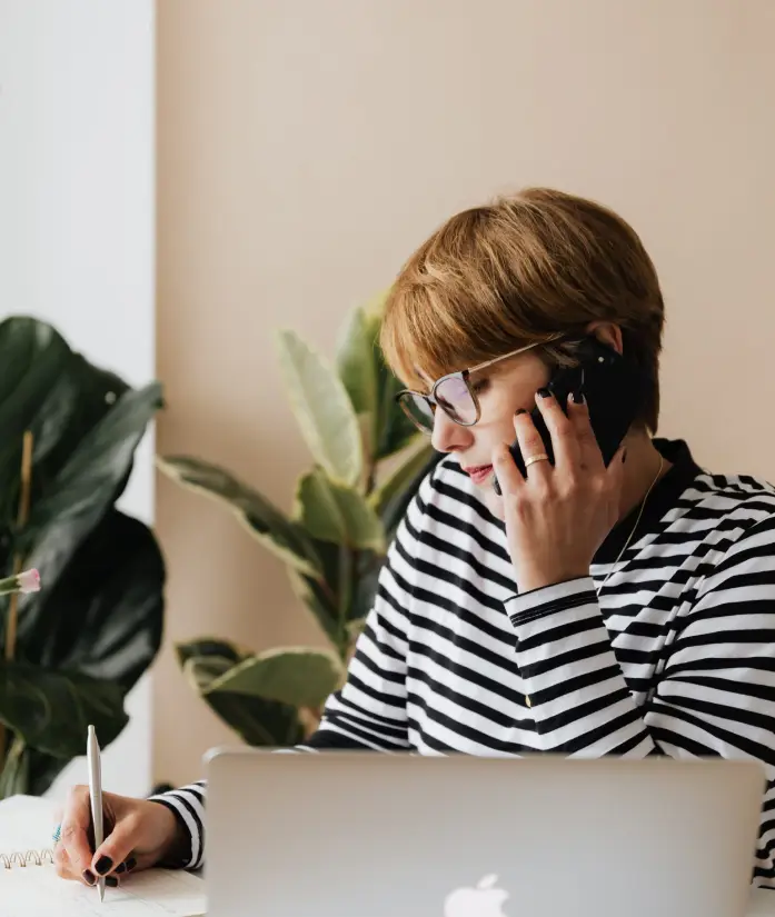 Woman talking on the phone while writing notes and working on a laptop.