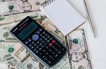 A calculator, notepad, and pen on top of dollar bills.